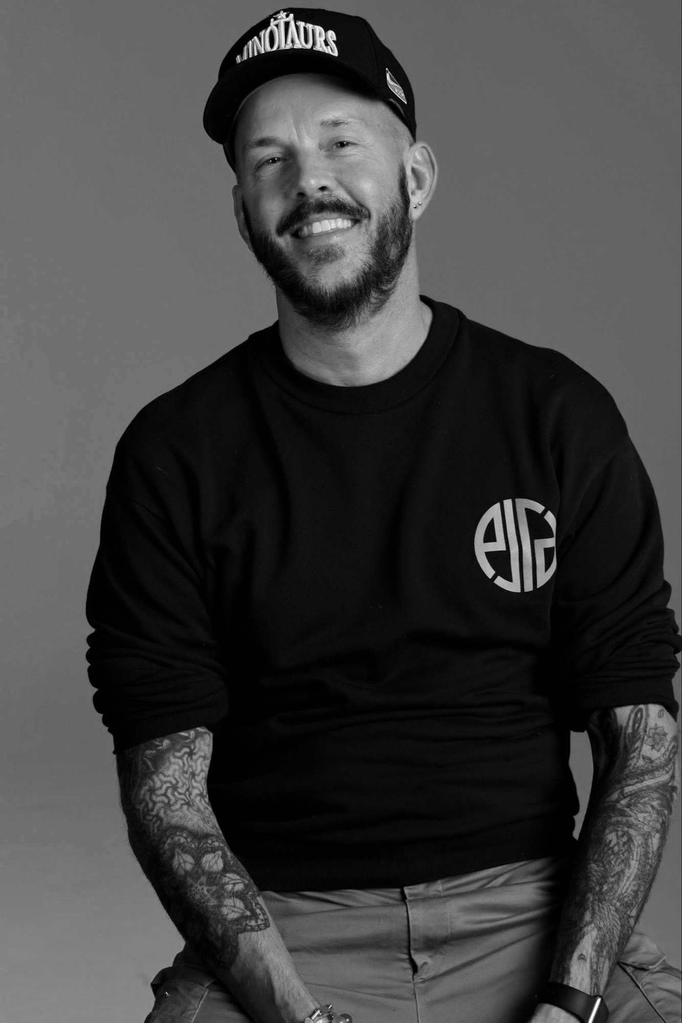 Portrait of Kevin Kollenda, black-and-white, seated, warm smile, black crewneck, tattoos on both arms.