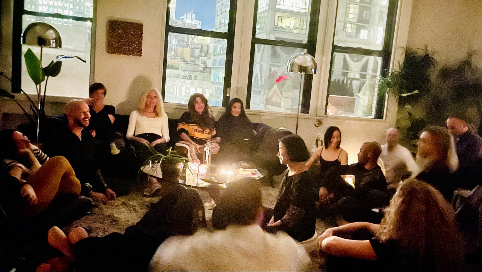 A gathering in a Manhattan loft at dusk, adults sitting in a circle around a low table of candles and books, the city skyline lit through tall windows behind them.
